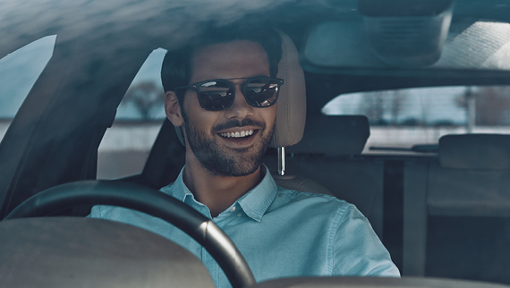 Man with sunglasses and a blue collared shirt smiling while sitting in a car.