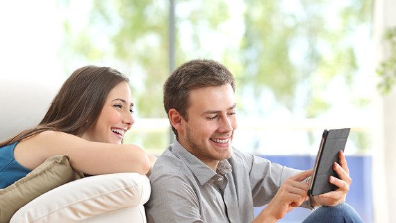 Couple sitting on a couch, smiling while looking at a tablet device.